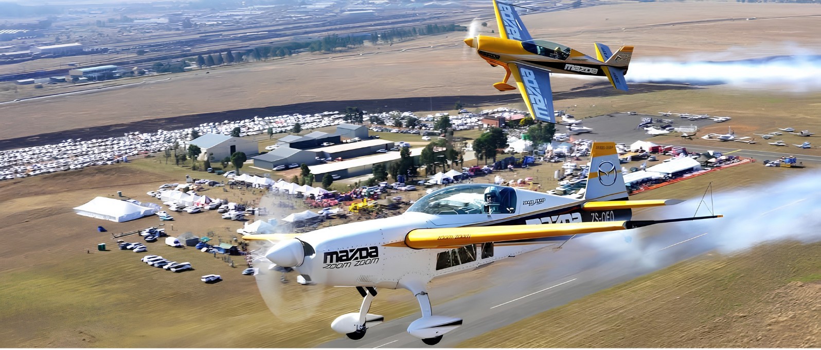 Two Mazda-liveried aerobatic planes trailing smoke low over Newcastle airfield, the crowd, tents and parked cars below