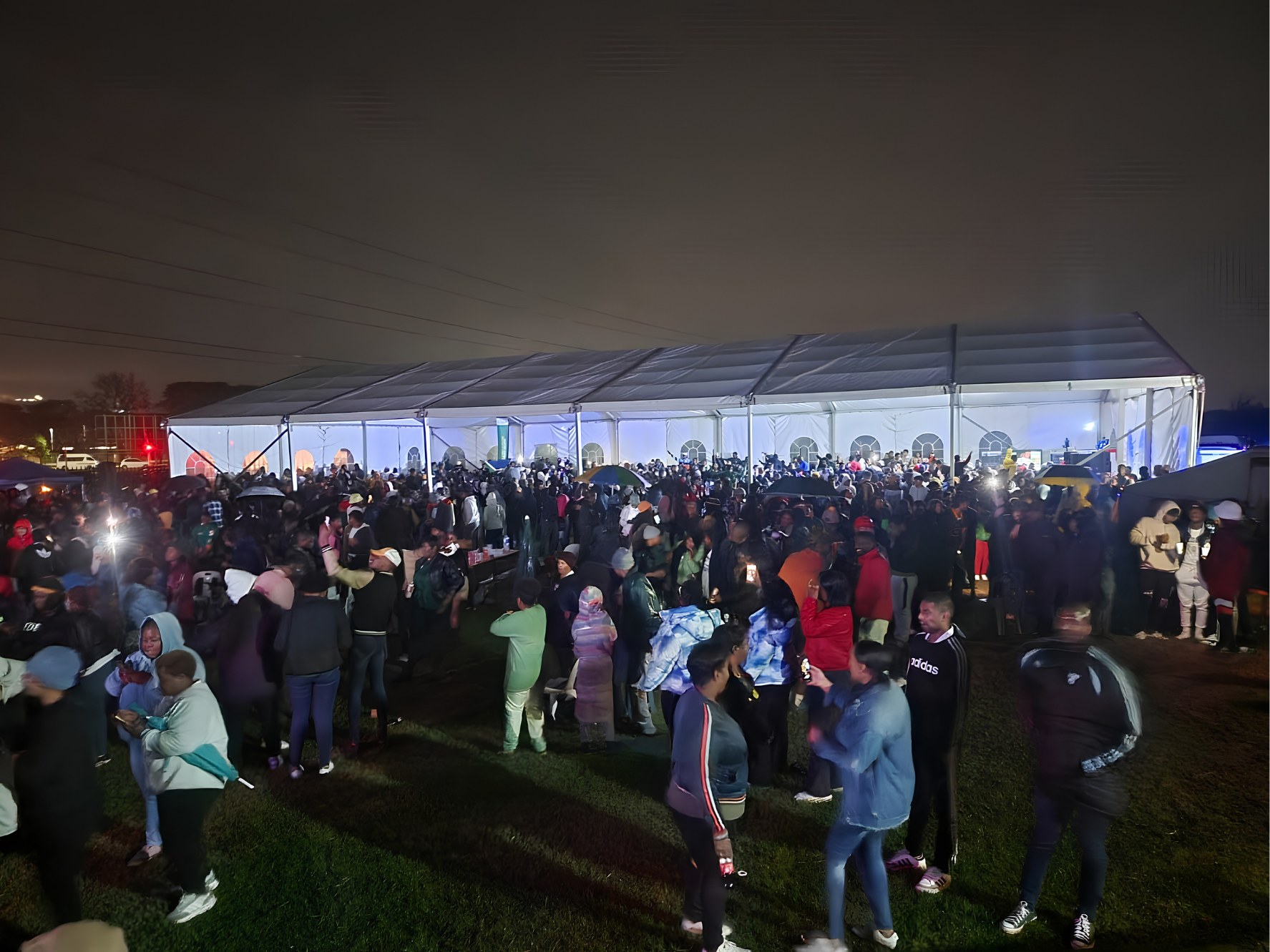 A big crowd gathered at night outside a lit marquee, umbrellas up in the drizzle