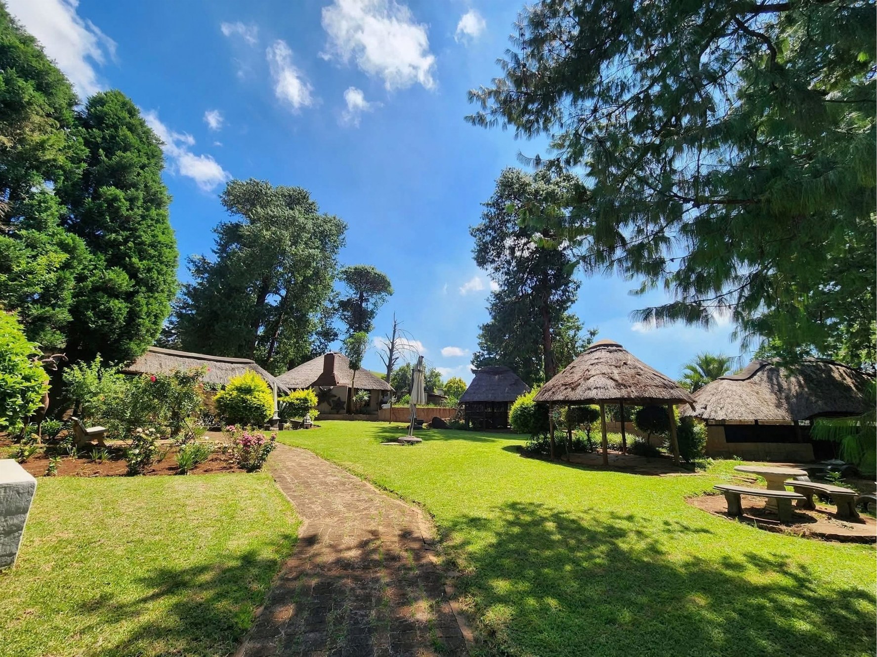 The Garden at The Pines: a brick path across sunny lawns to thatched gazebos among the trees