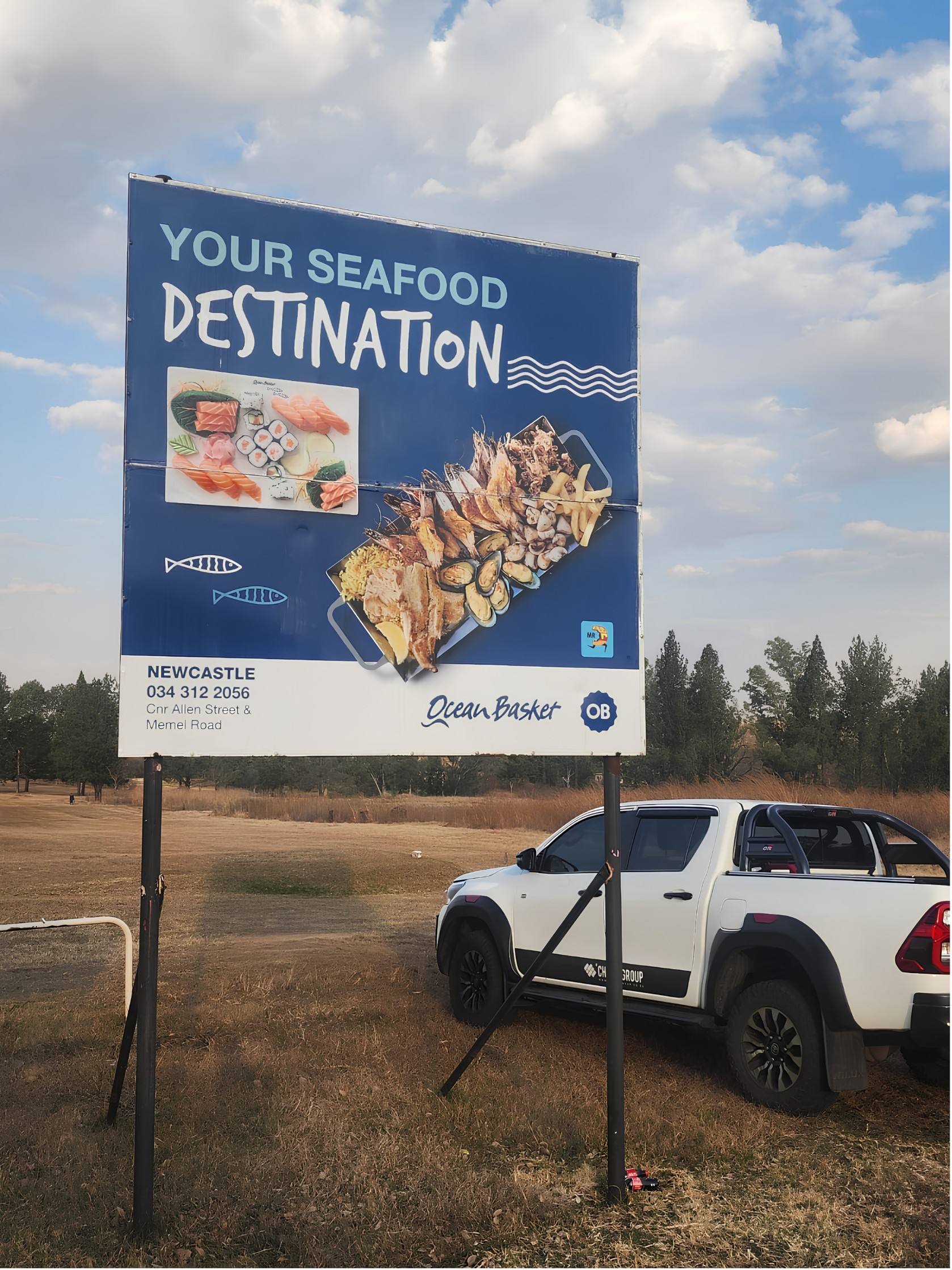 An Ocean Basket Newcastle billboard by The Print Lab beside a field, a Champ Group bakkie parked below